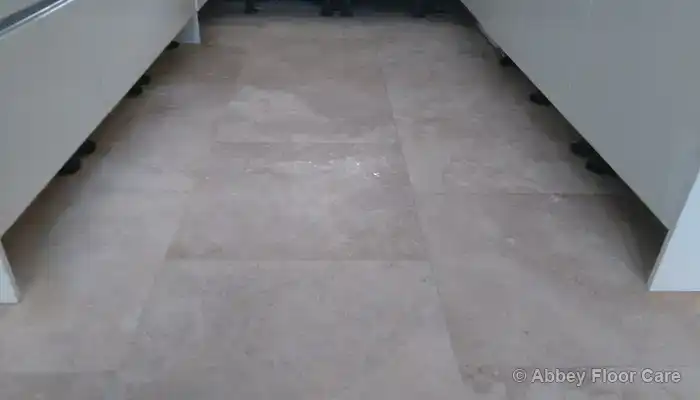 Worn travertine floor near kitchen units with dulled surface and uneven finish.